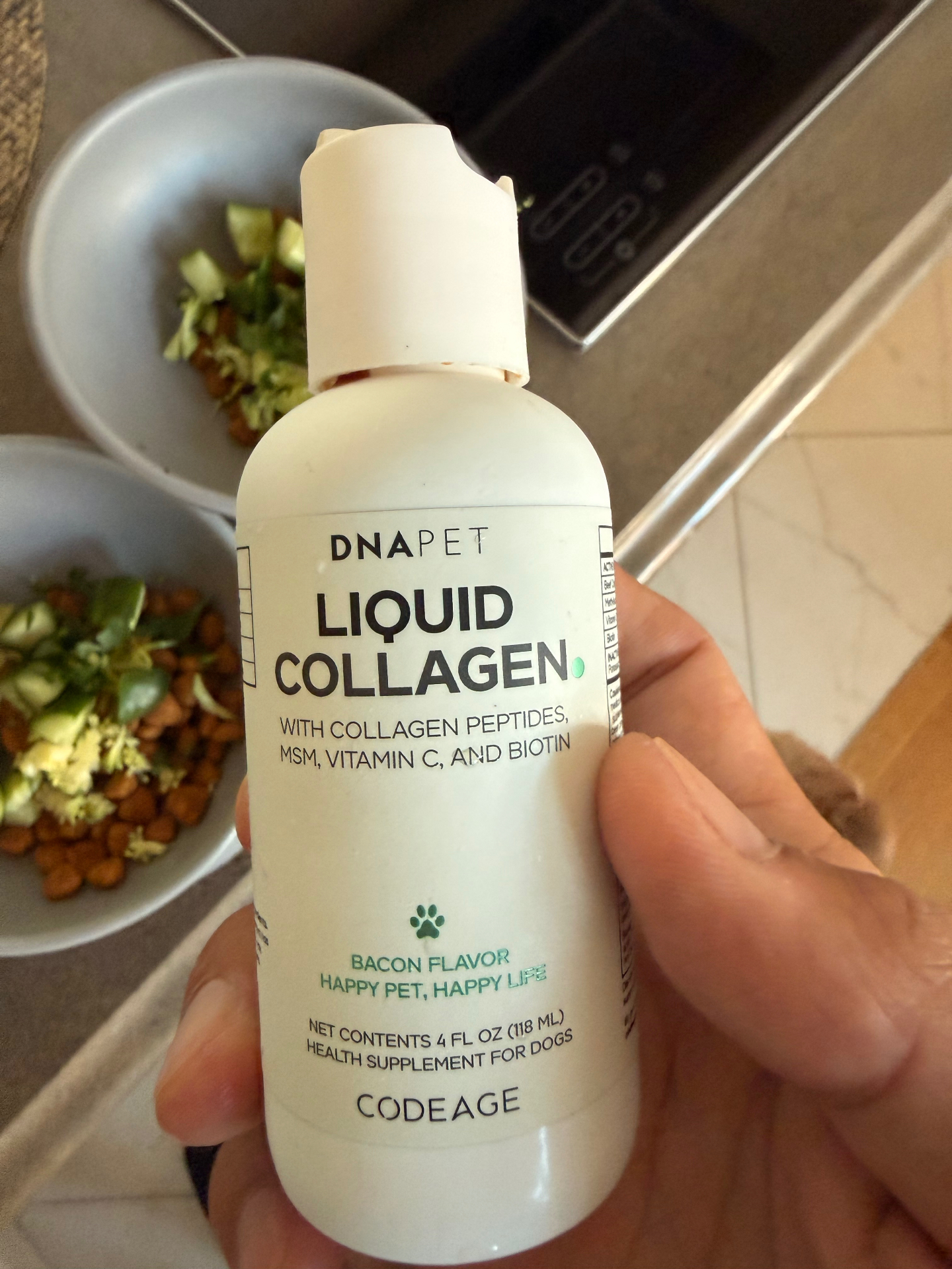 A hand holding a bottle of DNAPet liquid collagen supplement on a kitchen counter beside bowls of fresh dog food